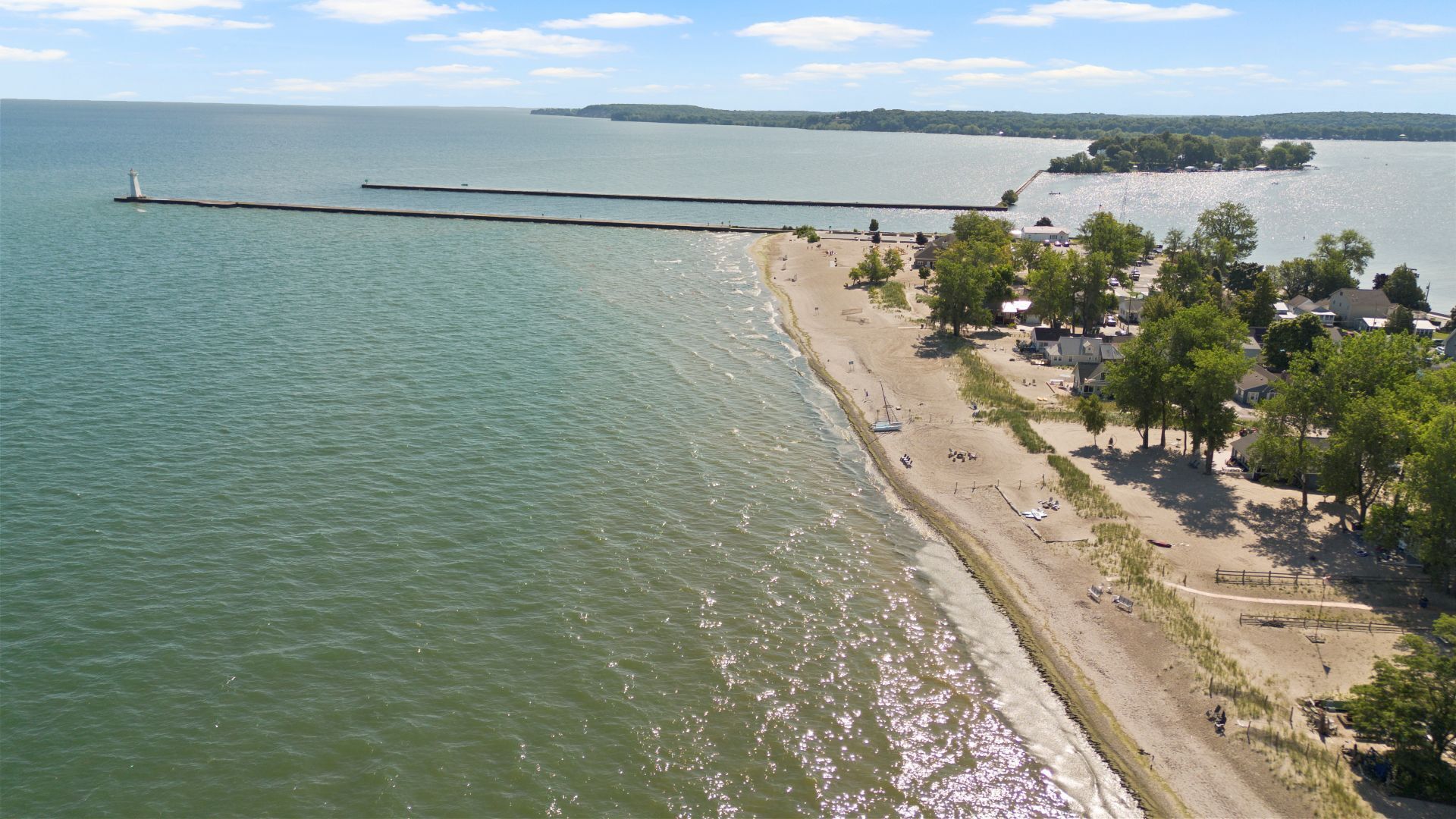 Une zone côtière avec une jetée qui s’avance dans l’eau, une plage avec des personnes et des arbres, et une côte lointaine parsemée de bâtiments.