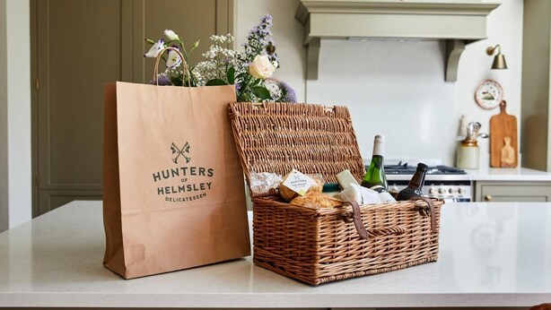Ein Korbkörbchen mit einer Weinflasche und einer braunen Papiertüte, beschriftet mit 'Hunters of Helmsley Delicatessen', auf einer Küchenarbeitsplatte.