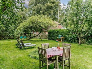 Um jardim com mesa e cadeiras de madeira, churrasqueira e uma casa ao fundo.