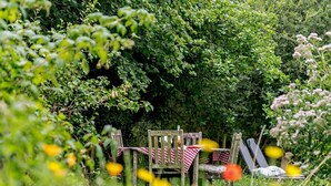 Een rustieke buitenruimte met een tafel en stoelen, omgeven door weelderig groen en bloemen.