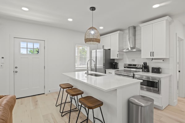 Une cuisine moderne avec des meubles blancs, un îlot central doté de tabourets, une hotte de cuisine en acier inoxydable et un luminaire suspendu.