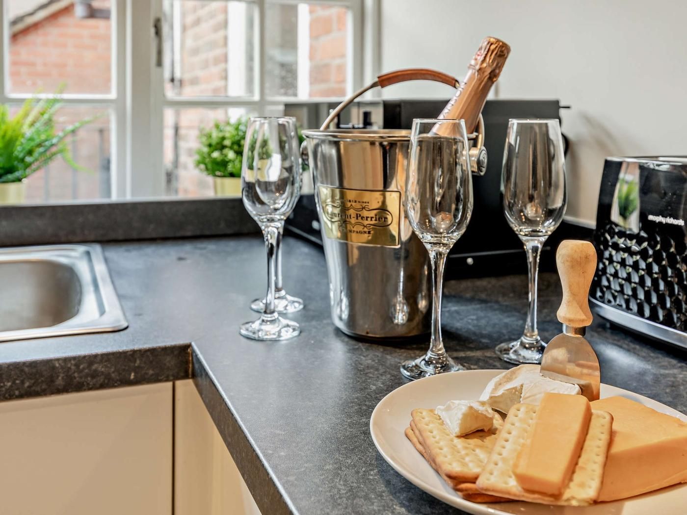 A kitchen counter with a bottle of champagne, glasses, and a cheese cutting board with cheese and crackers.