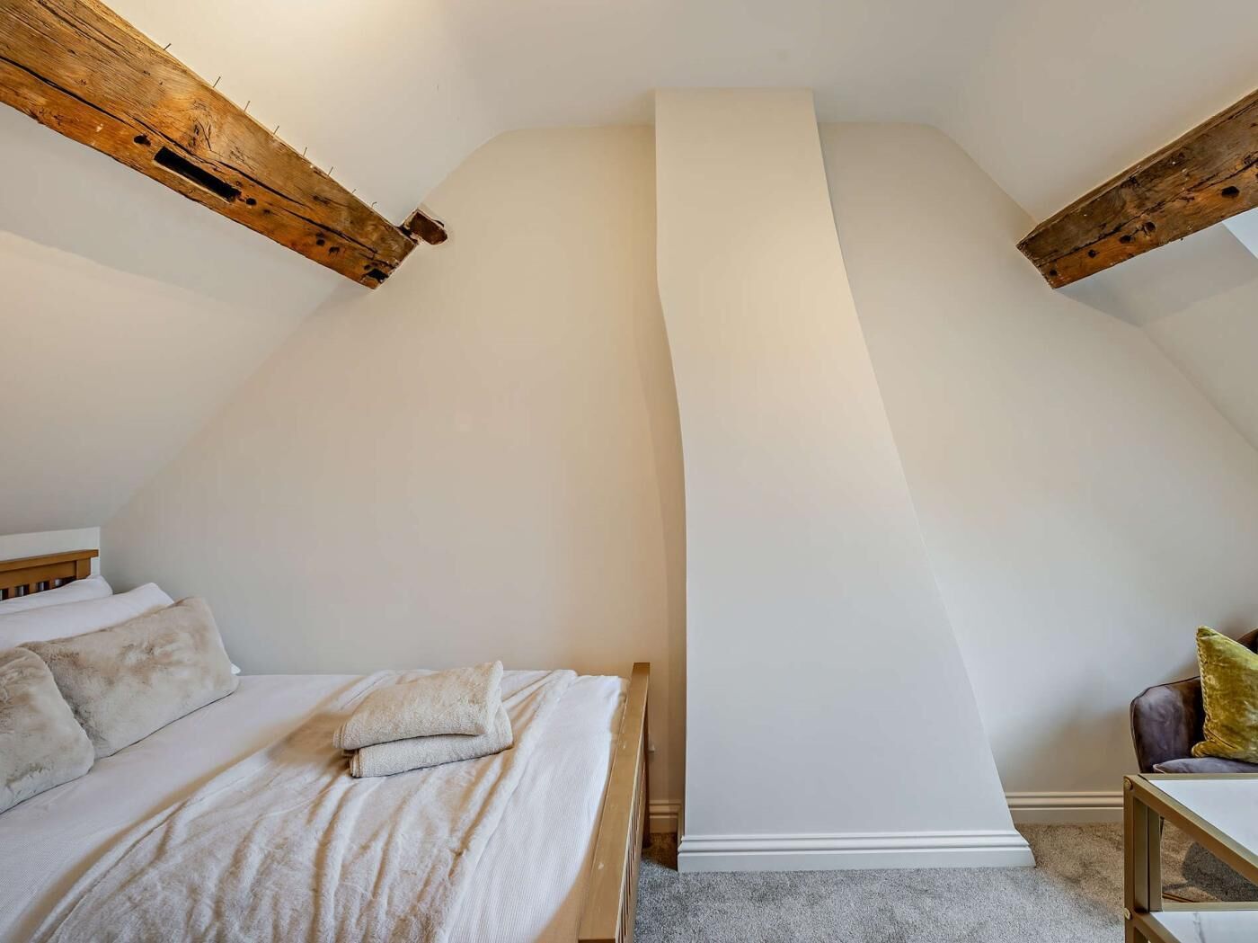 Een slaapkamer met een schuine kap, een bed met witte beddengoed, en een klein tafeltje met een kussen.