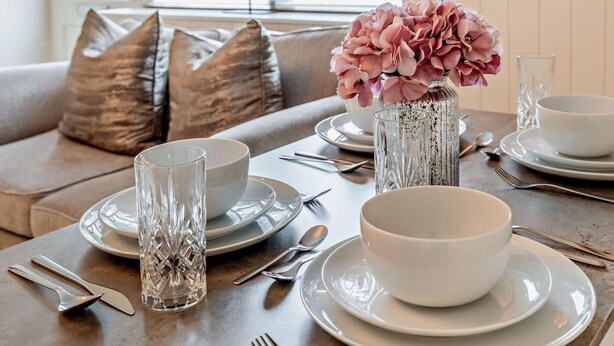 Une table à manger dressée avec des assiettes, des bols, des couverts et un vase rempli de fleurs.