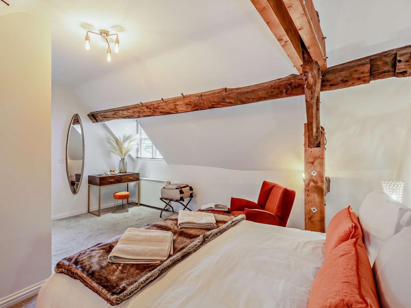 Une chambre avec un plafond en pente, une structure en poutres apparentes, un bureau avec une chaise, un lit avec des oreillers et un grand miroir.