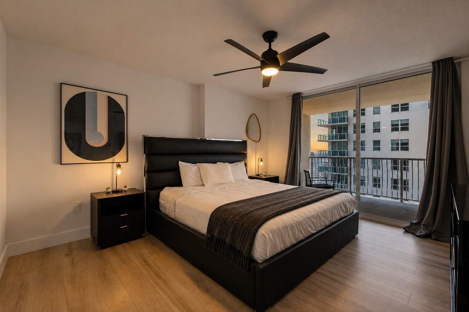 Un dormitorio moderno con una cama grande, un ventilador de techo, vistas de un edificio a través de la ventana y una obra de arte enmarcada en la pared.