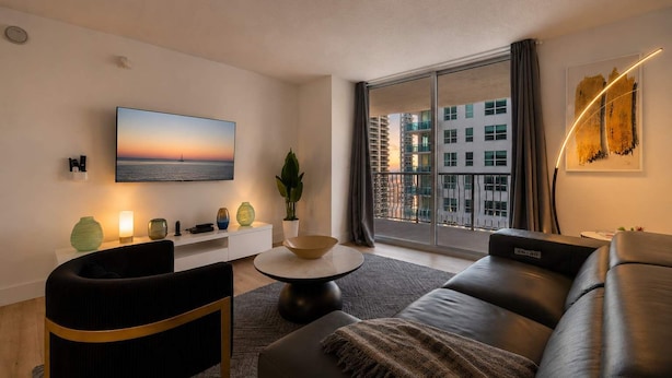 A modern living room with a large flat-screen TV, a sofa, a coffee table, and a view of a cityscape through a large window. - Luxury Brickell With City Views & Rooftop Pool (Miami)