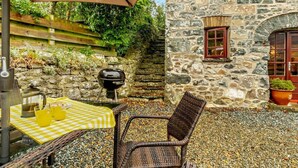 A stone building with a window and a door, a grill, a table with a checkered tablecloth, and a wicker chair. - 3 Bed in Haverfordwest (oc-92675) (Solva)