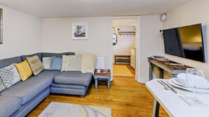 Un salon moderne avec un canapé gris, un téléviseur à écran plat, une table à manger avec des couverts, et un couloir menant à une chambre.