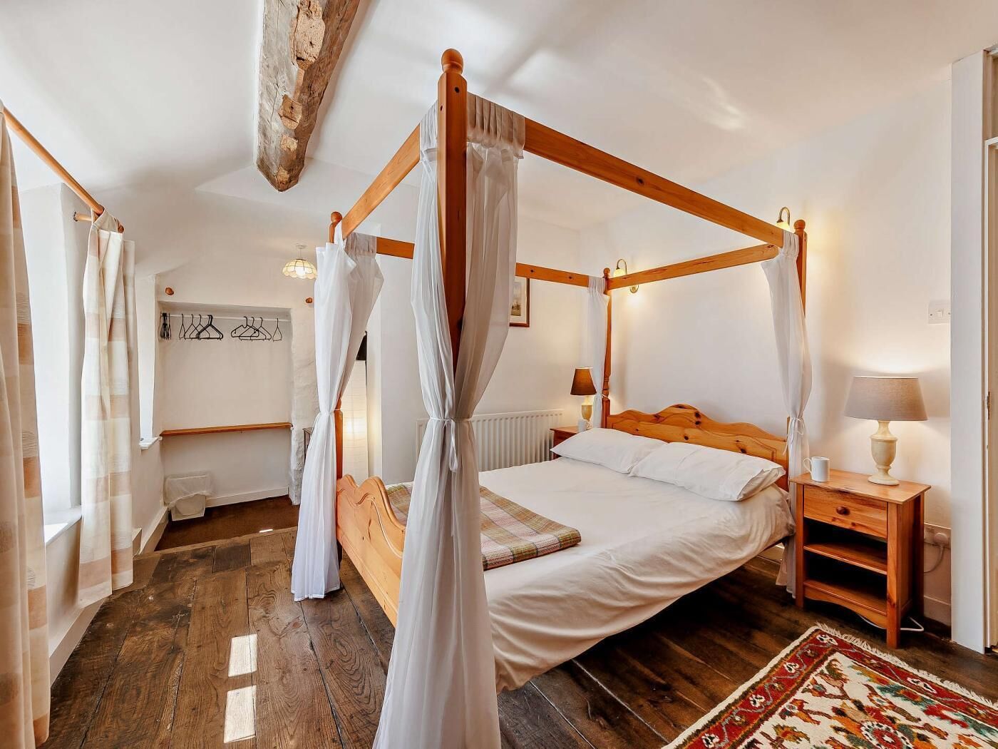A four-poster bed with white linens, a wooden nightstand with a lamp, and a patterned rug on a wooden floor.