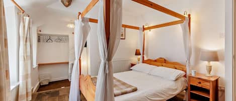 A four-poster bed with white linens, a wooden nightstand with a lamp, and a patterned rug on a wooden floor.