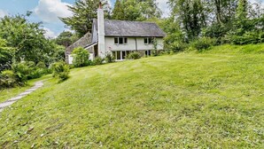 A two-story house with a stone chimney, surrounded by lush greenery and a sloped lawn. - 3 Bed in Machynlleth (oc-93083) (Cwm-Llinau)