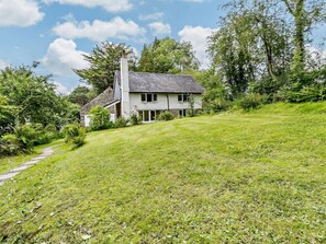 A two-story house with a stone chimney, surrounded by lush greenery and a sloped lawn. - 3 Bed in Machynlleth (oc-93083) (Cwm-Llinau)