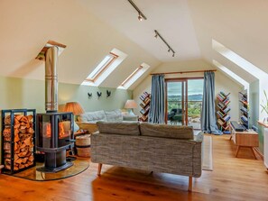 A cozy loft bedroom with a built-in wood stove, a comfortable sofa, a large window with a view, and a balcony with bookshelves. - 3 Bed in Gairloch (oc-92714) (Badachro)