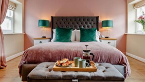 Un dormitorio con cabecera capitonada, dos mesitas de noche con lámparas, cama con una combinación de colores rosa y verde, un banco con artículos para el desayuno y una ventana con cortinas.