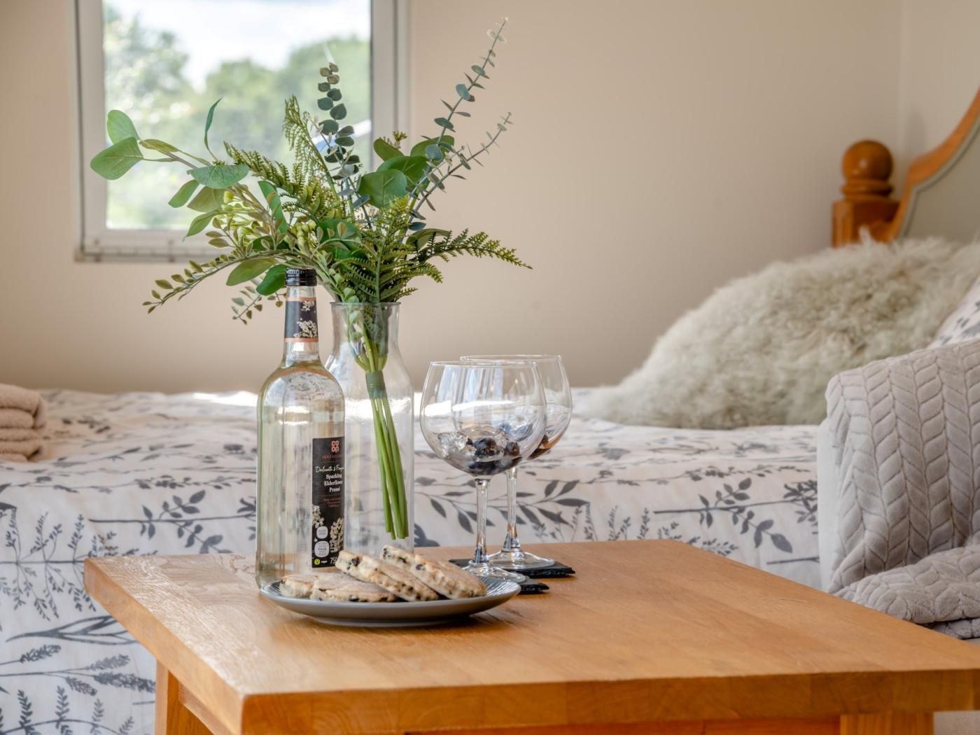 A bedroom with a bed, a bedside table with a bottle, a vase with greenery, and a glass.