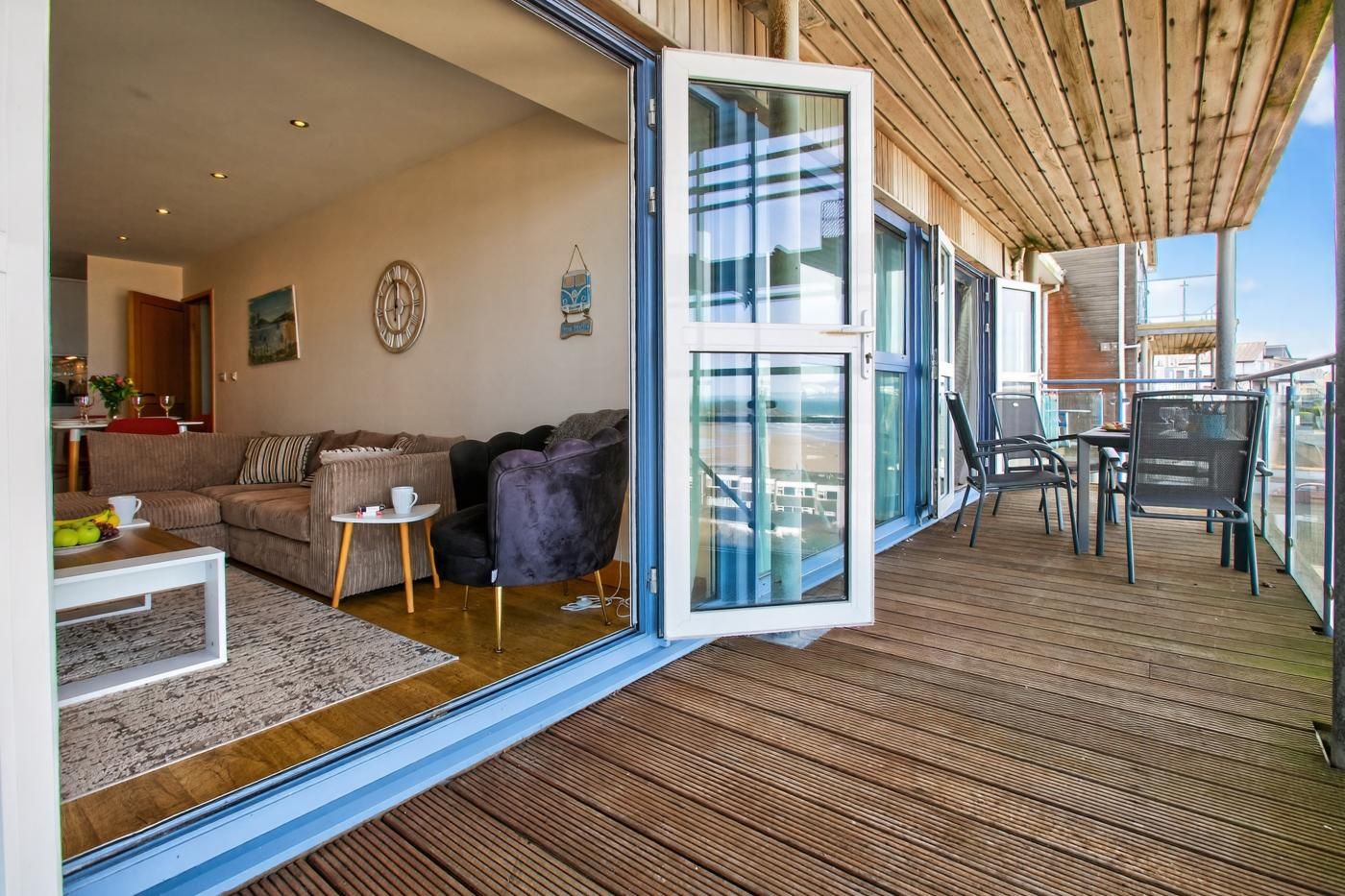 Ein modernes Wohnzimmer mit Sofa, Sessel und Couchtisch, ein Balkon mit Sitzgelegenheit im Freien und eine große Schiebeglas-Tür.