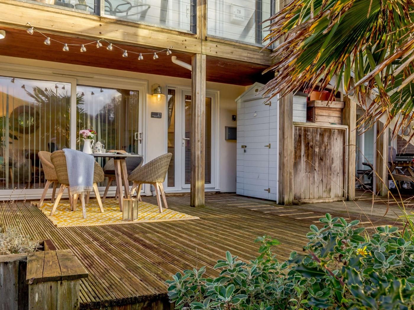 Um deck de madeira com mesa e cadeiras, uma porta de vidro deslizante e uma pequena área de jardim.