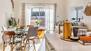 Een moderne woonkamer met een glazen eettafel, rieten stoelen, een salontafel en een bank. Er is een groot raam met gordijnen, een plant en een macramé wanddecoratie.