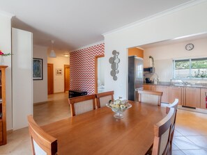 Ein Essbereich mit Holztisch, Stühlen, einer Schüssel mit Lebensmitteln, einer Küche mit Kühlschrank und einer Uhr an der Wand.