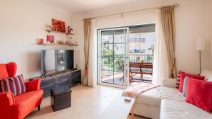 Ein Wohnzimmer mit Fernseher, Sofa und Sesseln, ein Balkon mit Tisch und Stühlen sowie Blick auf Gebäude und Grünflächen.