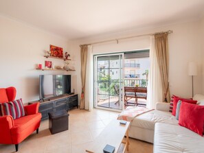 A living room with a TV, sofa, and chairs, a balcony with a table and chairs, and a view of buildings and greenery. - Apt. Luz da Manhã, with sea views (Luz)