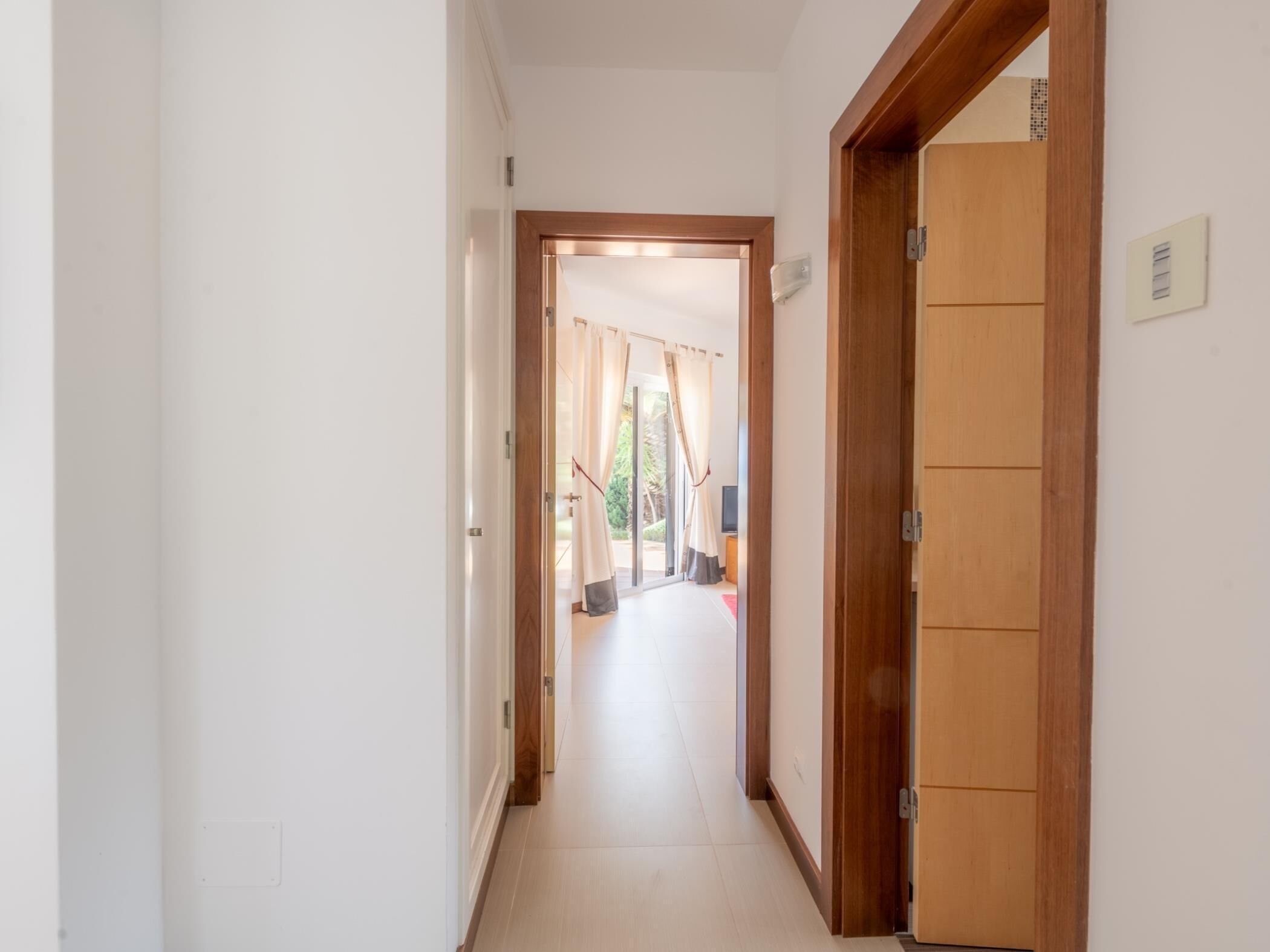 Un pasillo con una puerta de madera a la derecha, una pared blanca y vista a una habitación con cortina.
