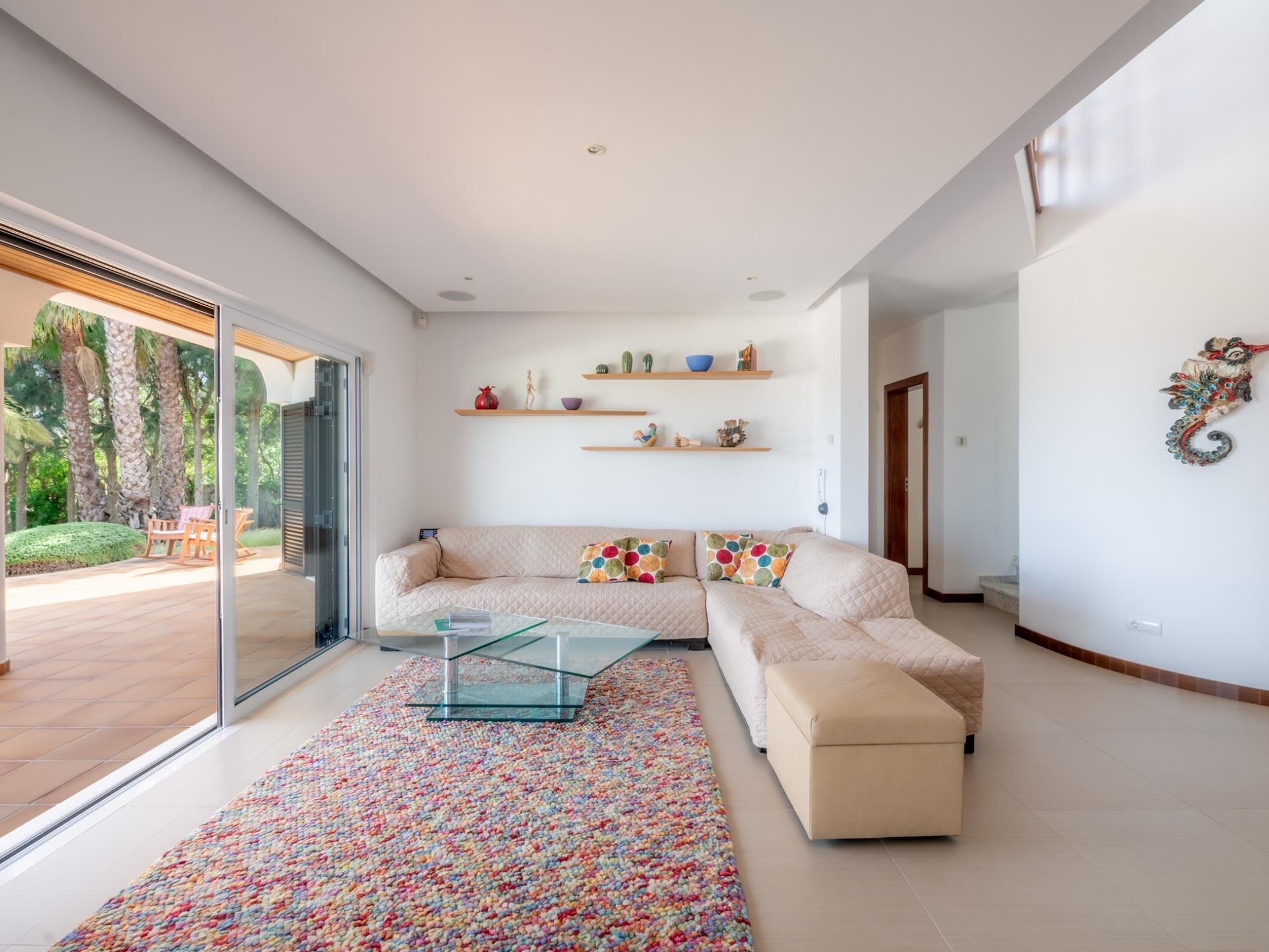 Ein modernes Wohnzimmer mit Glastür zum Patio, einem Sofa mit bunten Kissen, einem Glastisch und dekorativen Regalen.