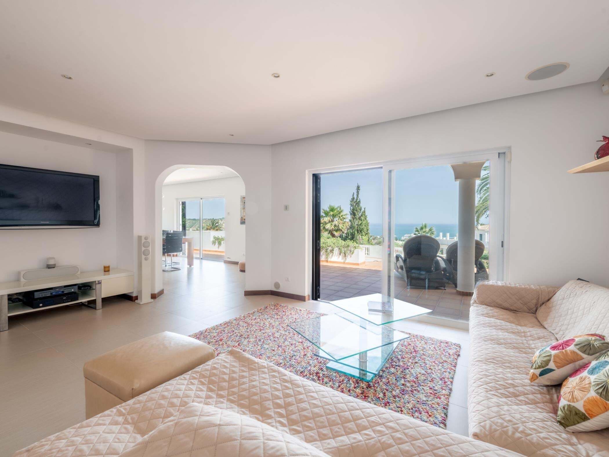 A modern living room with a large TV, a glass coffee table, and a sofa. There is a balcony with a view of the sea.