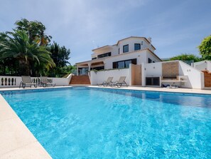 Un espace moderne aménagé au bord de la piscine, avec une piscine à l’eau cristalline, des chaises longues et une maison à deux étages dotée d’un balcon.