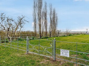 Zona vallada para que jueguen los perros, con un letrero, una puerta de metal y árboles al fondo.