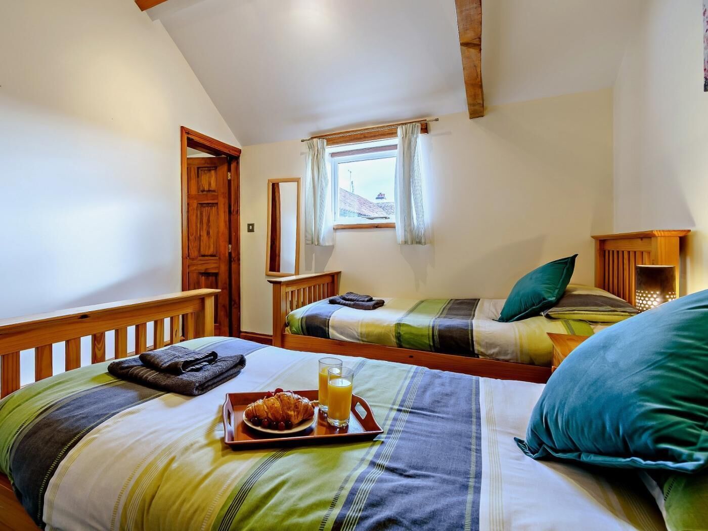 A bedroom with two beds, a window with curtains, a wooden door, and a tray with croissants and juice.