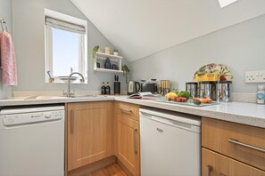 A modern kitchen with a dishwasher, wooden cabinets, a window, and various kitchen appliances. - 1 Bed in Sturminster Newton (oc-93841) (Hazelbury Bryan)