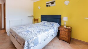 A bedroom with a bed, a nightstand, a lamp, a chair, a shelf, and a painting on the wall. - Villa Sea La Vie | Exclusive Villa with Private Pool near Palmares Golf Course (Vale da Lama - Odiáxere)