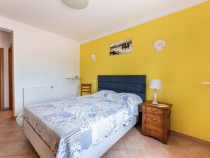 A bedroom with a bed, a nightstand, a lamp, a chair, a shelf, and a painting on the wall. - Villa Sea La Vie | Exclusive Villa with Private Pool near Palmares Golf Course (Vale da Lama - Odiáxere)