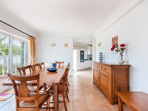 A dining area with a wooden table and chairs, a kitchen in the background, and a view through a large window. - Villa Sea La Vie | Exclusive Villa with Private Pool near Palmares Golf Course (Vale da Lama - Odiáxere)