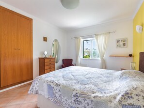 Une chambre avec un grand lit, une armoire en bois, une commode, un miroir, une fenêtre avec des rideaux et une étagère fixée au mur.