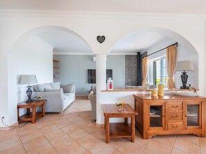 A living room with a sofa, a wooden table, and a cabinet. There is a kitchen area with a refrigerator and a window with curtains. - Villa Sea La Vie | Exclusive Villa with Private Pool near Palmares Golf Course (Vale da Lama - Odiáxere)