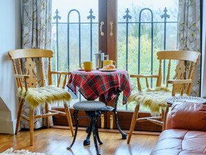A room with two wooden chairs, a small table with a plaid tablecloth, and a view through a window with a metal gate. - 4 Bed in Stroud (oc-93690) (Burcombe)