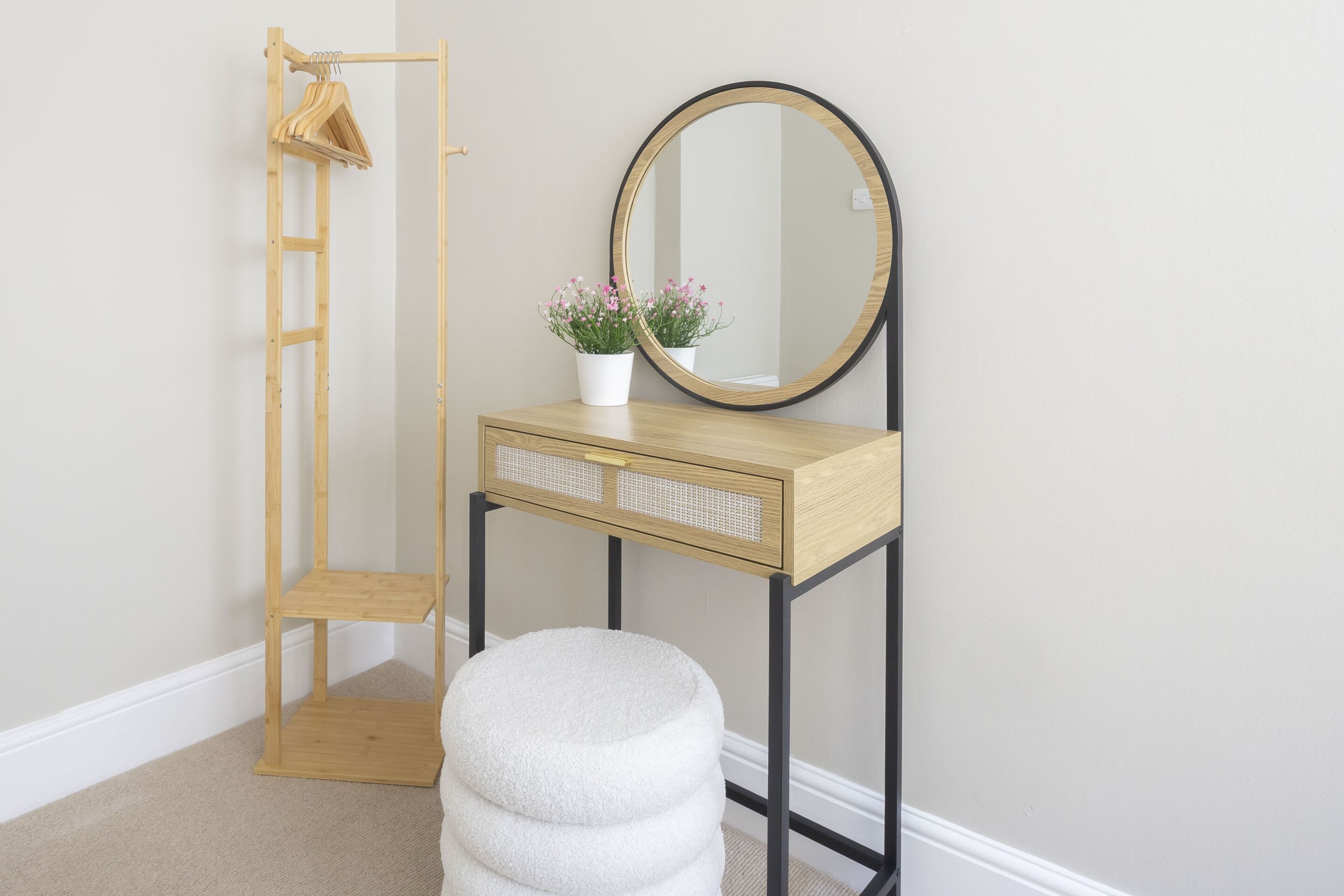 A room with a wooden vanity, a round mirror, a small stool, a wooden hanger rack, and a potted plant.
