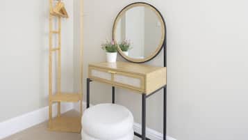 A room with a wooden vanity, a round mirror, a small stool, a wooden hanger rack, and a potted plant.