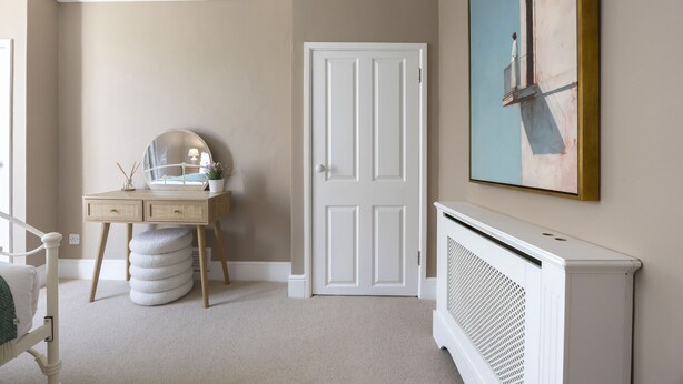 Et værelse med en hvid dør, et skrivebord med et spejl, en radiator og et billede på væggen.