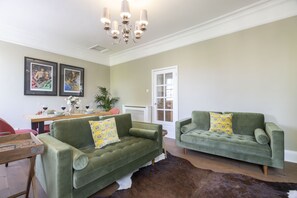 Een woonkamer met twee groene banken, een eettafel met stoelen, kunstwerken in lijsten aan de muur en een kroonluchter.