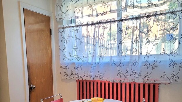 A room with a round table, two chairs, a radiator, a door, and a window with a floral curtain.