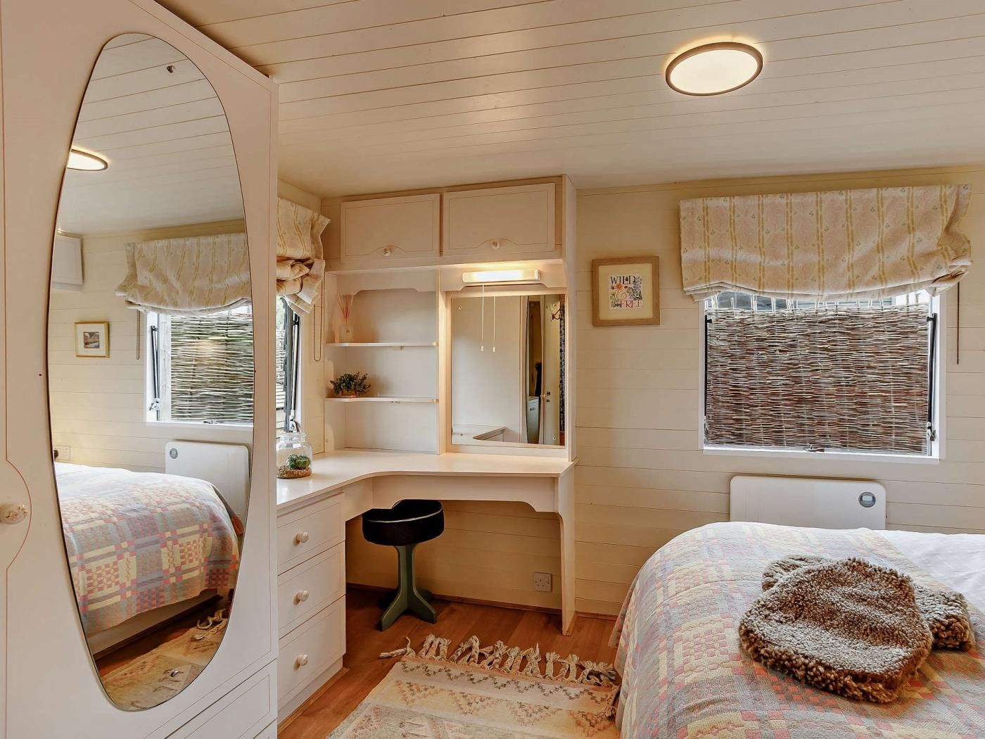 A bedroom with a large mirror, a desk, a window with curtains, and a bed with a patterned bedspread.