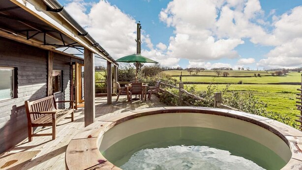 A wooden deck with a hot tub, a table with chairs, and a view of a green field.