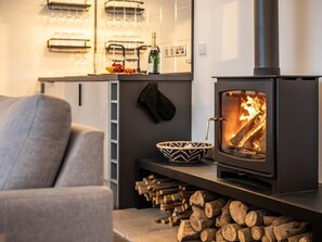 A modern living room with a wood-burning stove, a grey sofa, a black and white woven bowl, and neatly stacked firewood. - 5 Bed in Fowey (oc-93849) (Polruan)