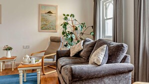 Ein Wohnzimmer mit grauem Sofa, einem Holztisch, einem kleinen Beistelltisch, einer Topfpflanze und einem Bild an der Wand.