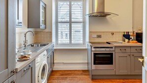 A compact kitchen with a washing machine, oven, and a window with blinds. - 1 Bed in Marazion (oc-53251) (Marazion)