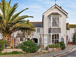 A white building with a bay window and a small balcony, surrounded by greenery and a cobblestone path. - 1 Bed in Marazion (oc-53251) (Marazion)